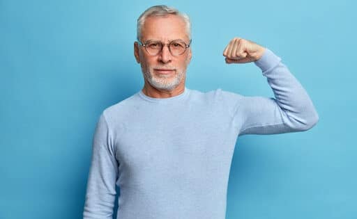 Homem branco, idoso, de suéter azul claro, com o fundo azul e o braço flexionado como forma de força, para simbolizar o Dia Mundial de Combate ao Câncer de Próstata