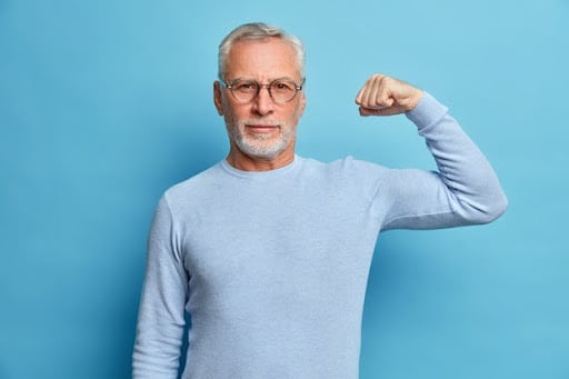 Homem branco, idoso, de suéter azul claro, com o fundo azul e o braço flexionado como forma de força, para simbolizar o Dia Mundial de Combate ao Câncer de Próstata